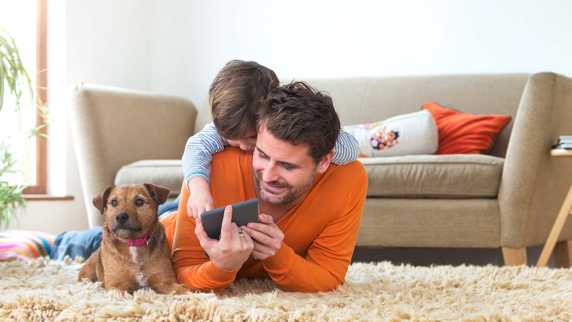 Man laying down on rug. Looking at his phone with toddler and dog included.