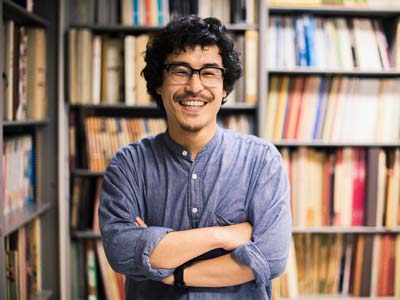 Young man with glasses smiling arms folded in front of a library bookshelf
