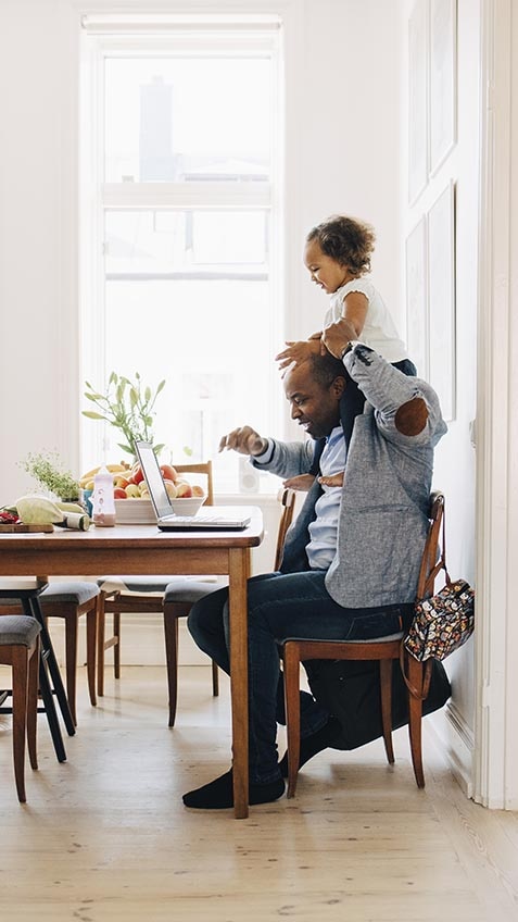 Man working from home on computer, with a toddler on his shoulders.